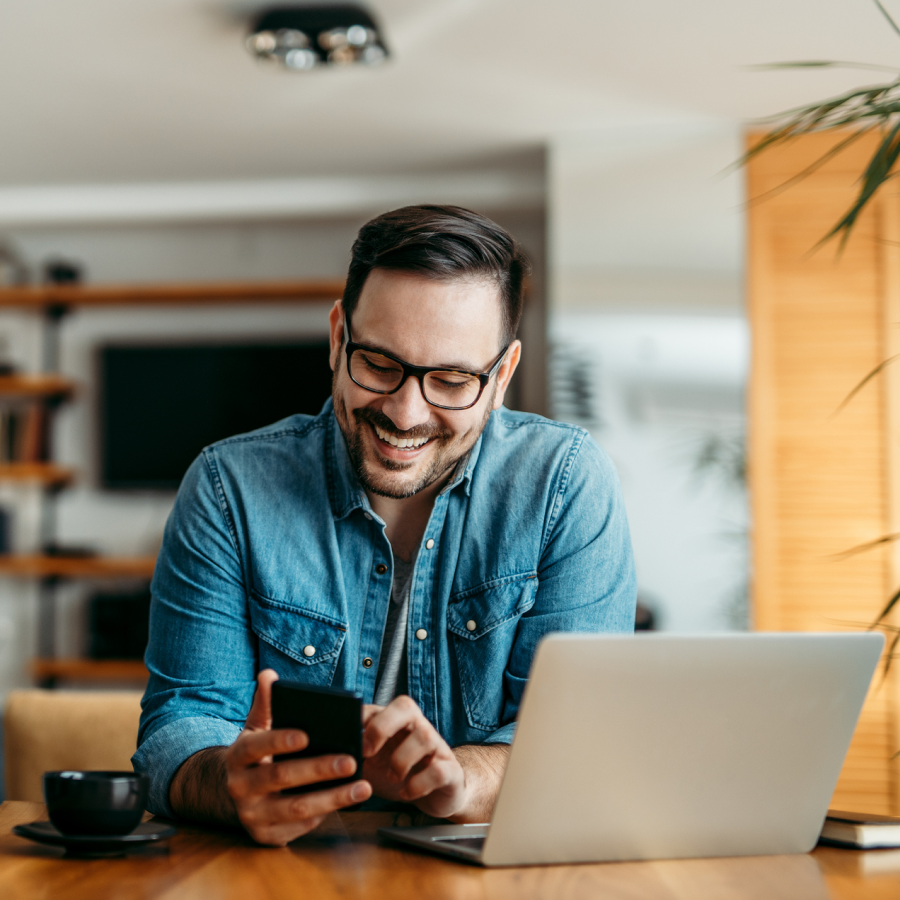 Muž sedí pri stole, v ruke drží mobilný telefón a pred sebou má otvorený notebook. Vizualizácia služby Business Asset, ktorá pomáha firmám spravovať svoj majetok digitálne a bezpečne.