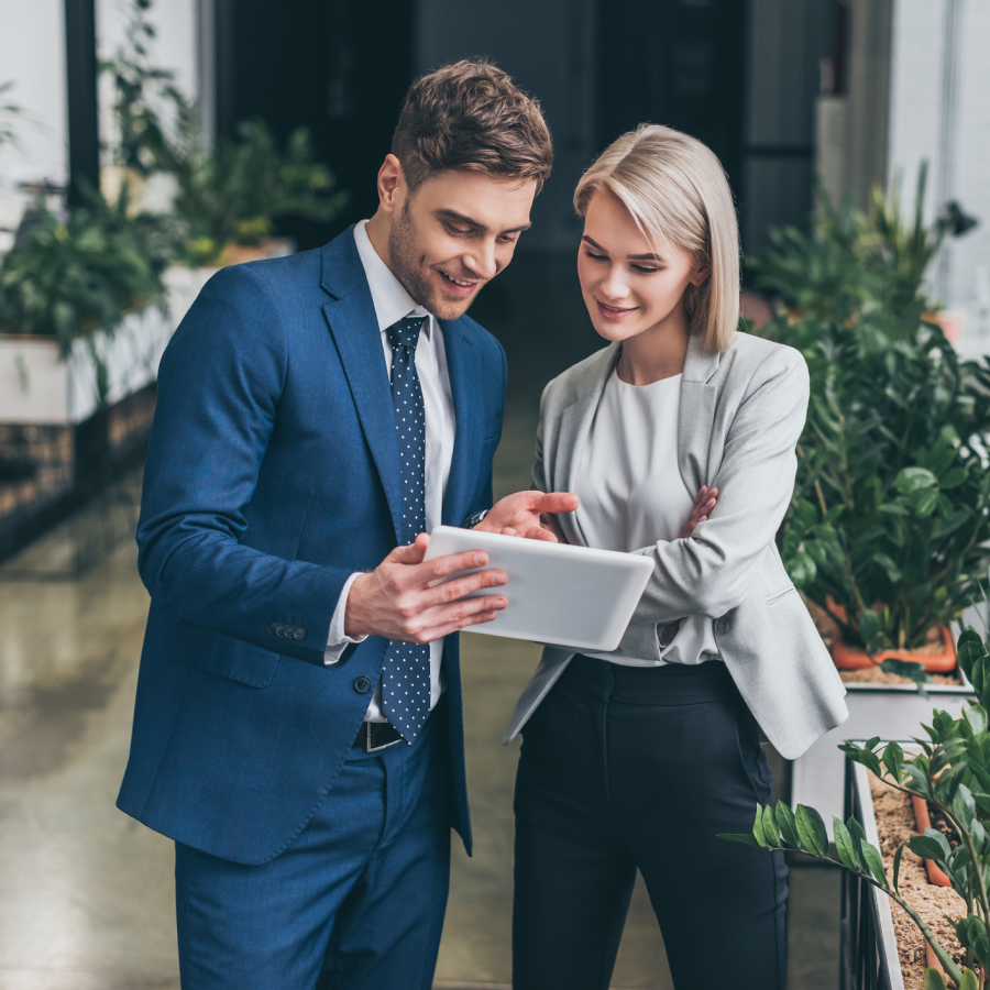 Muž v obleku a žena v biznis outfite stoja v kancelárii a spolu sa pozerajú na tablet. Vizualizácia profesionálneho poradenstva a výhody služby Telekom Business Asset pre firmy.