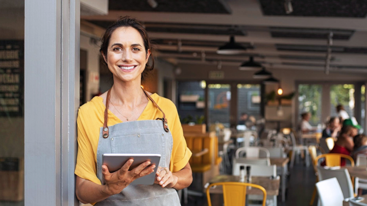 Usmievavá žena v zástere drží tablet pred reštauráciou – znázorňuje prehľad o majetku v gastro segmente s viacerými prevádzkami.
