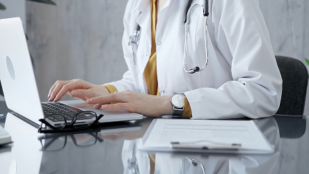 Lekárka v bielom plášti pracuje na notebooku pri stole s dokumentáciou. Vizualizácia výhod služby Telekom Cloud Server pre zdravotníctvo bez potreby riešiť infraštruktúru.