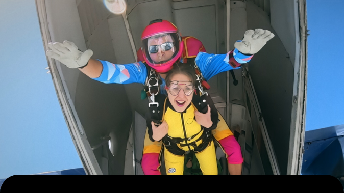 Tandemový zoskok – inštruktor s prilbou a okuliarmi stojí s pripútanou ženou v otvorených dverách lietadla pred skokom. Symbol dôvery trust kampan 2026