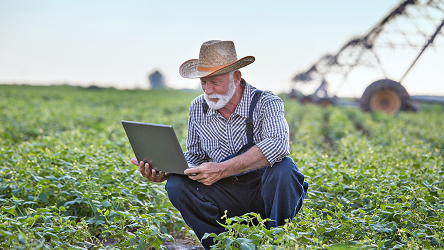 Poľnohospodár v klobúku pracuje s tabletom na poli. Ilustruje IoT konektivitu a využitie smart senzorov v poľnohospodárstve.