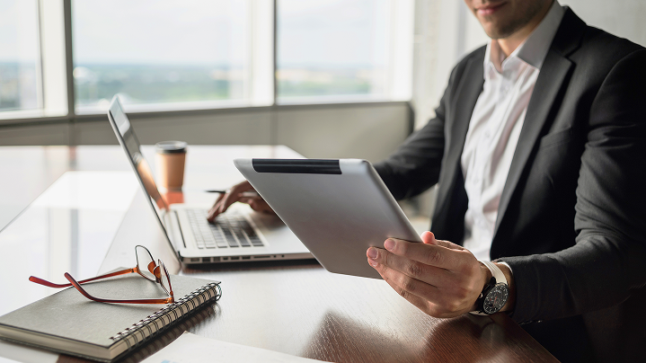 Muž v obleku pracuje s tabletom pri notebooku v kancelárii.