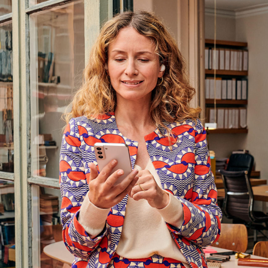 Žena v pestrofarebnom saku drží v ruke smartfón a prezerá si SMS správy. Symbolizuje efektívnu zákaznícku komunikáciu pomocou Business Messaging riešení.