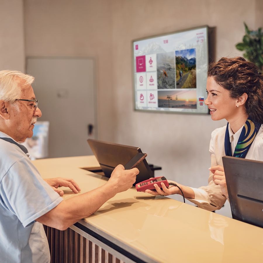 Recepčná s úsmevom podáva zákazníkovi platobný terminál – za nimi obrazovka s Telekom digitálnym obsahom. Ilustruje využitie digitálneho marketingového riešenia v praxi.