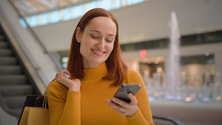 Mladá žena s nákupnými taškami a mobilom v ruke sa usmieva v nákupnom centre. Ilustruje rýchlu marketingovú komunikáciu so zákazníkmi v retaili.