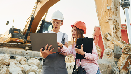 Dvojica pracovníkov na stavenisku s tabletmi pred stavebnými strojmi – monitoring vozidiel a techniky na zvýšenie efektivity v stavebníctve.