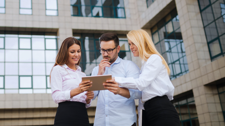 Tri osoby pred budovou sledujú tablet – symbol efektívneho nasadenia Smart Meteringu pri správe budov.